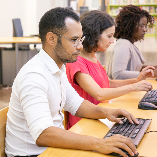 Adults of diverse backgrounds working on desktop computers in a library or classroom setting.