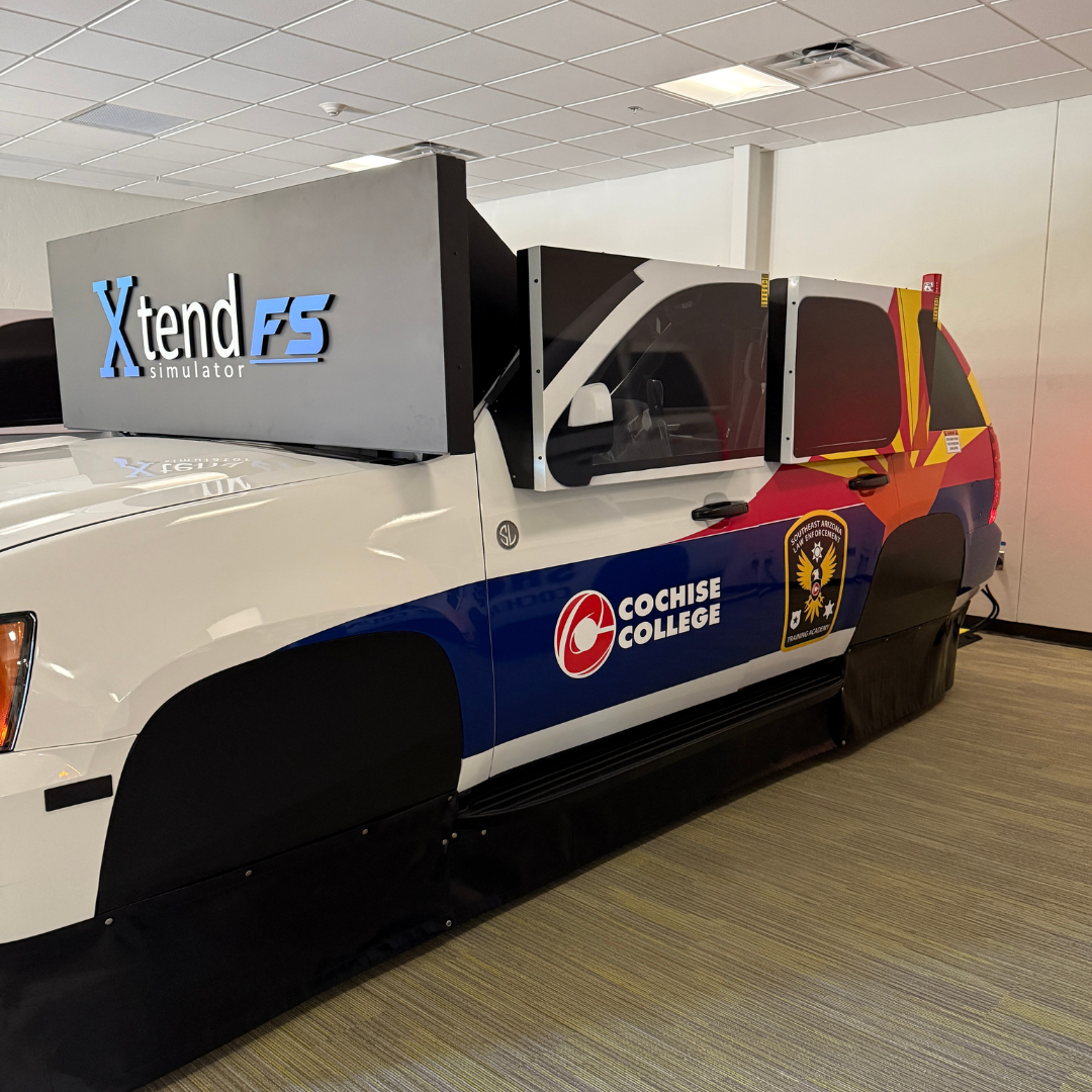 A white SUV-shaped driving simulator with Cochise College and state logos on the side, set up indoors.