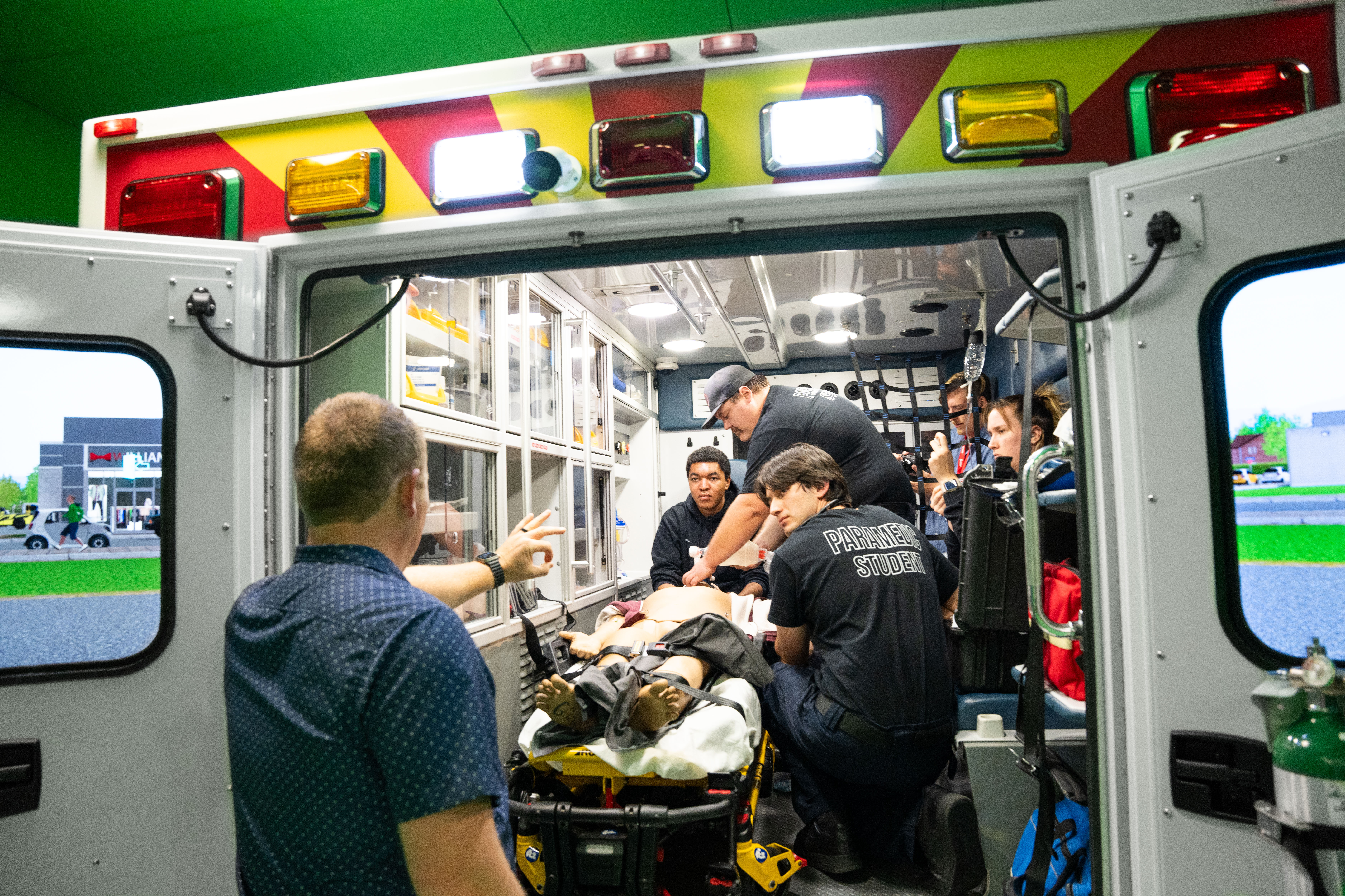 Paramedic students practice emergency care on a simulation mannequin inside an ambulance with an instructor guiding them.