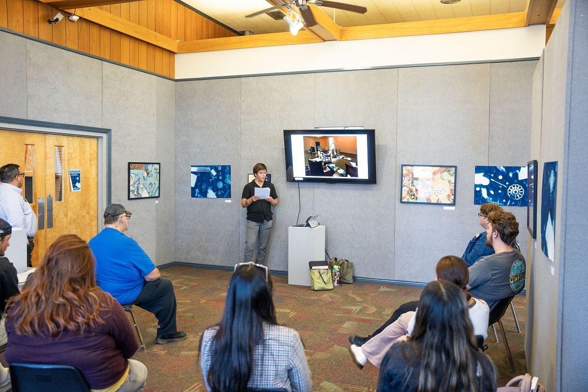 Speaker presenting at an art exhibition in a gallery, with audience seated and artwork displayed on the walls.