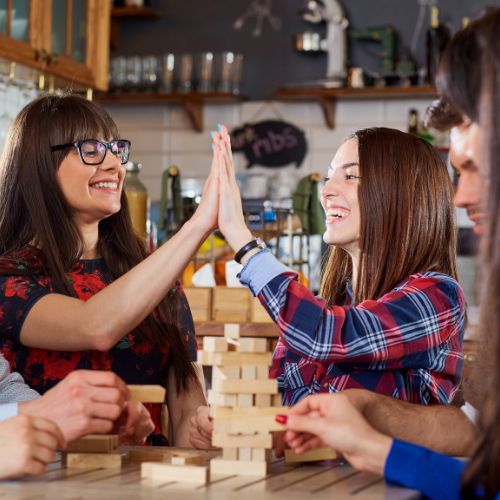 Group of young adults playing a block-stacking game together at a table, two women smiling and high-fiving in celebration.