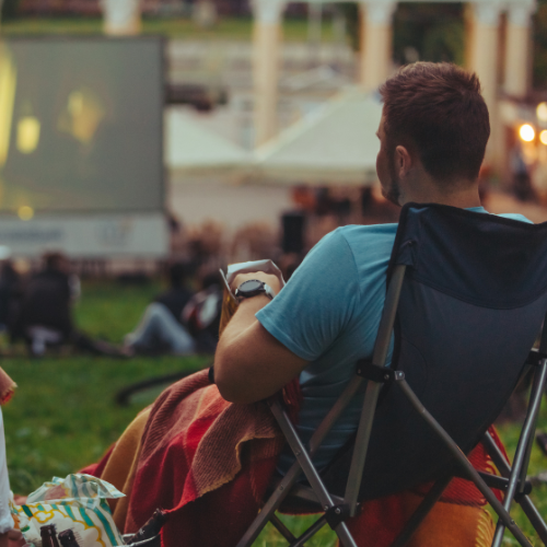 Man sitting in a lawn chair with a blanket watching an outdoor movie on a big screen.