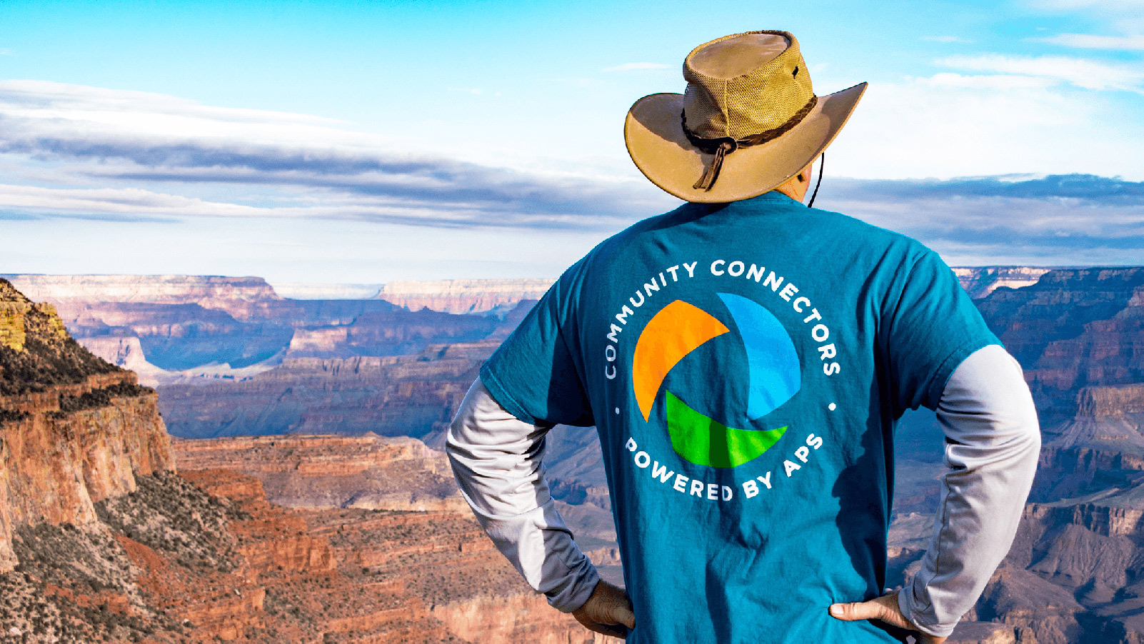 Person in a hat overlooking a canyon, wearing a teal “Community Connectors – Powered by APS” shirt.