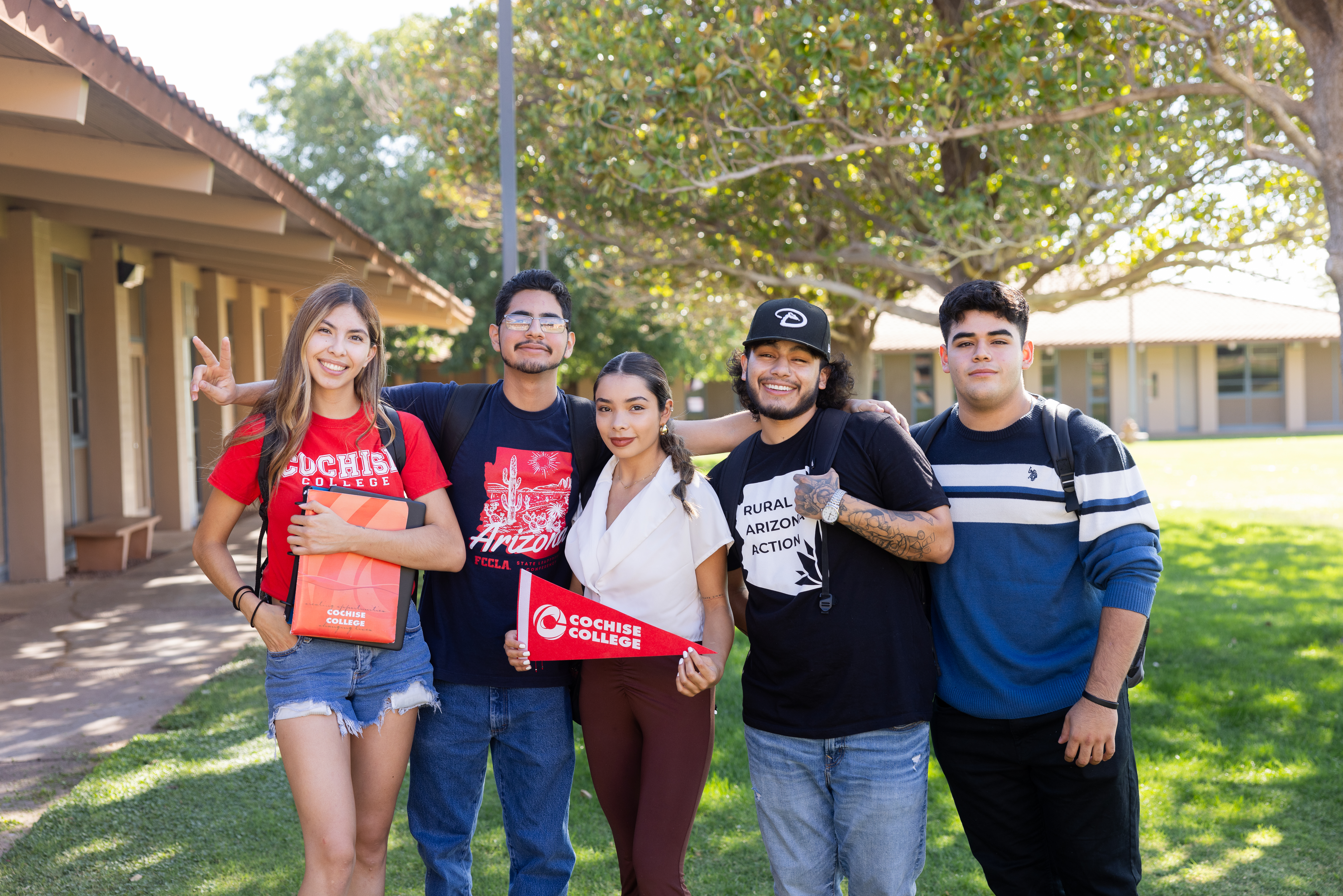 Five Cochise College students standing together outside on the Douglas campus, smiling and posing for the camera. One student holds a Cochise College pennant, and another flashes a peace sign. Trees and campus buildings are visible in the background.